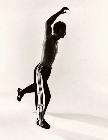 Monochromatic image of a muscular adult male posing in black and white track pants in a studio with a light background and dynamic lighting.