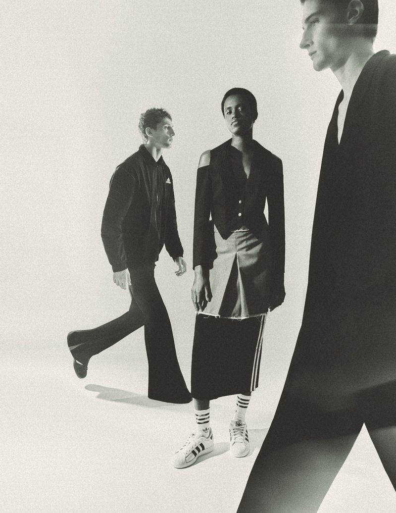 Three fashion models in a monochrome studio shot. A male and female model stand in the center, while a third man's torso is cropped.