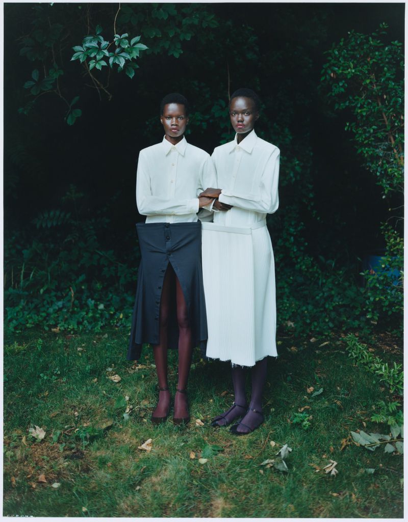 Two dark-skinned adult women stand in an outdoor setting; they're wearing collared shirts and skirts in a fashion editorial style.