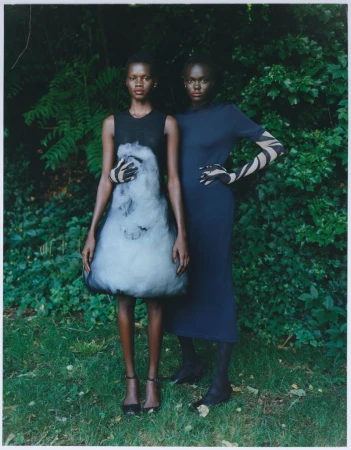 Two dark-skinned women stand in a garden setting, one in a black and white dress, the other in navy, gazing forward with neutral expressions.
