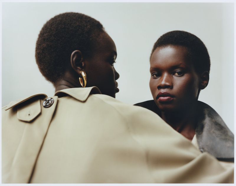 Two dark-skinned women in trench coats pose in a close-up studio shot. One woman faces the camera, the other is angled away with gold earrings.