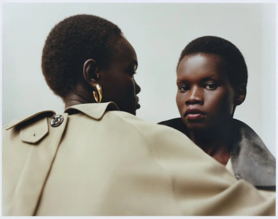 Two dark-skinned women in trench coats pose in a close-up studio shot. One woman faces the camera, the other is angled away with gold earrings.