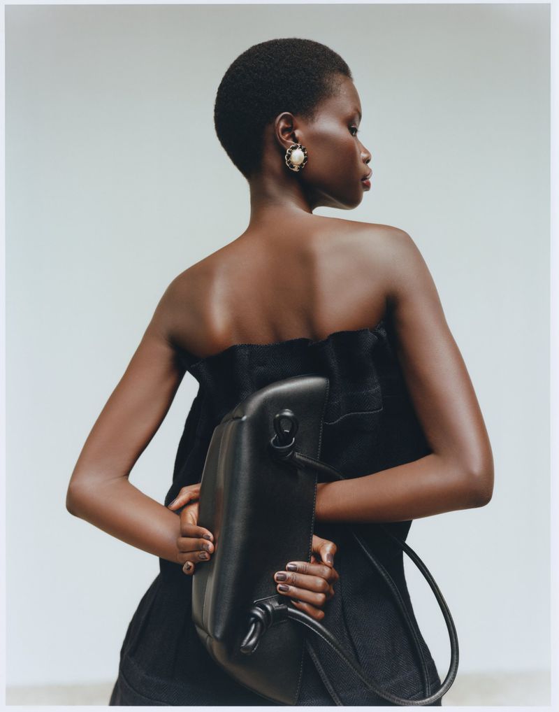 Back view of a black female model in a strapless black dress, adorned with pearl earrings and holding a black leather bag.
