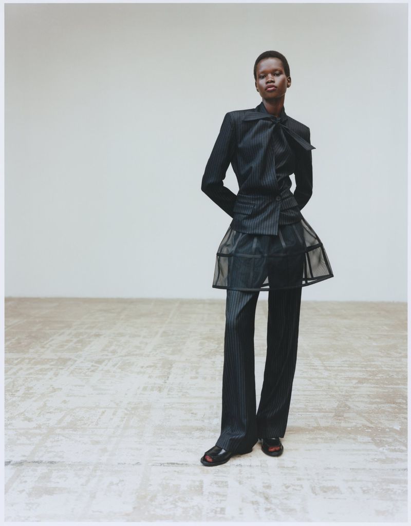 Fashion model in a pinstripe suit and black open-toe heels strikes a pose against a neutral wall in a fashion editorial shot.
