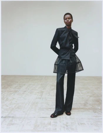 Fashion model in a pinstripe suit and black open-toe heels strikes a pose against a neutral wall in a fashion editorial shot.