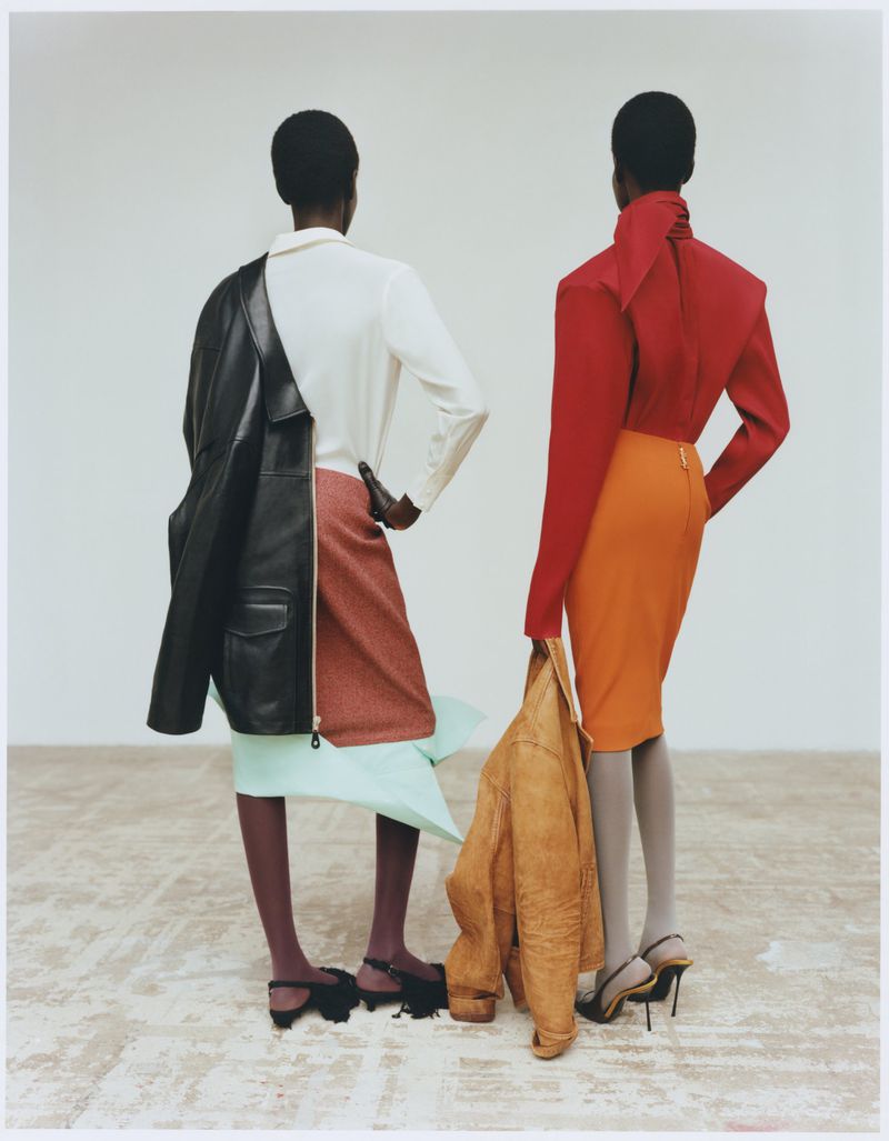 Two black female models in fashionable outfits stand in front of a white background, one with a black leather jacket on her shoulder.