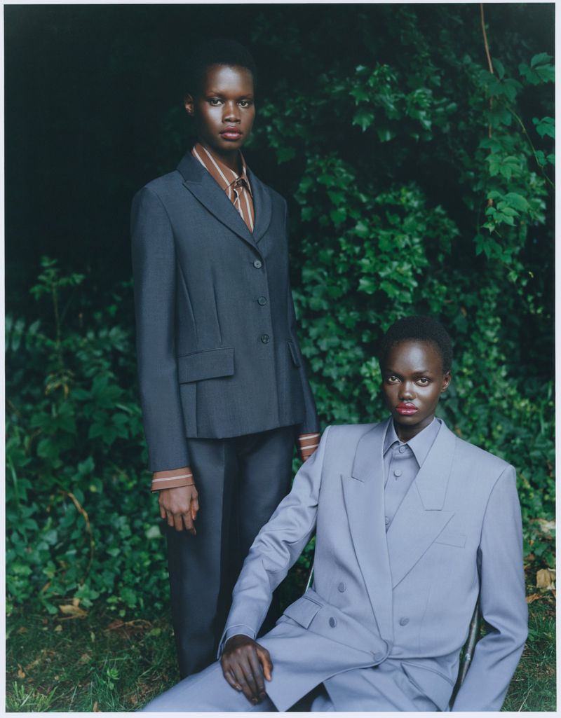Two women pose in tailored suits against a green foliage backdrop, showcasing modern fashion and natural lighting in a portrait orientation.
