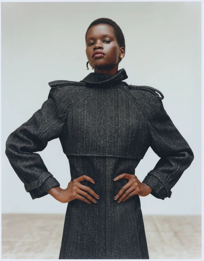 Model Achol Kuir wearing a dark structured coat with hands on hips against a plain light studio background for Harper's Bazaar France.