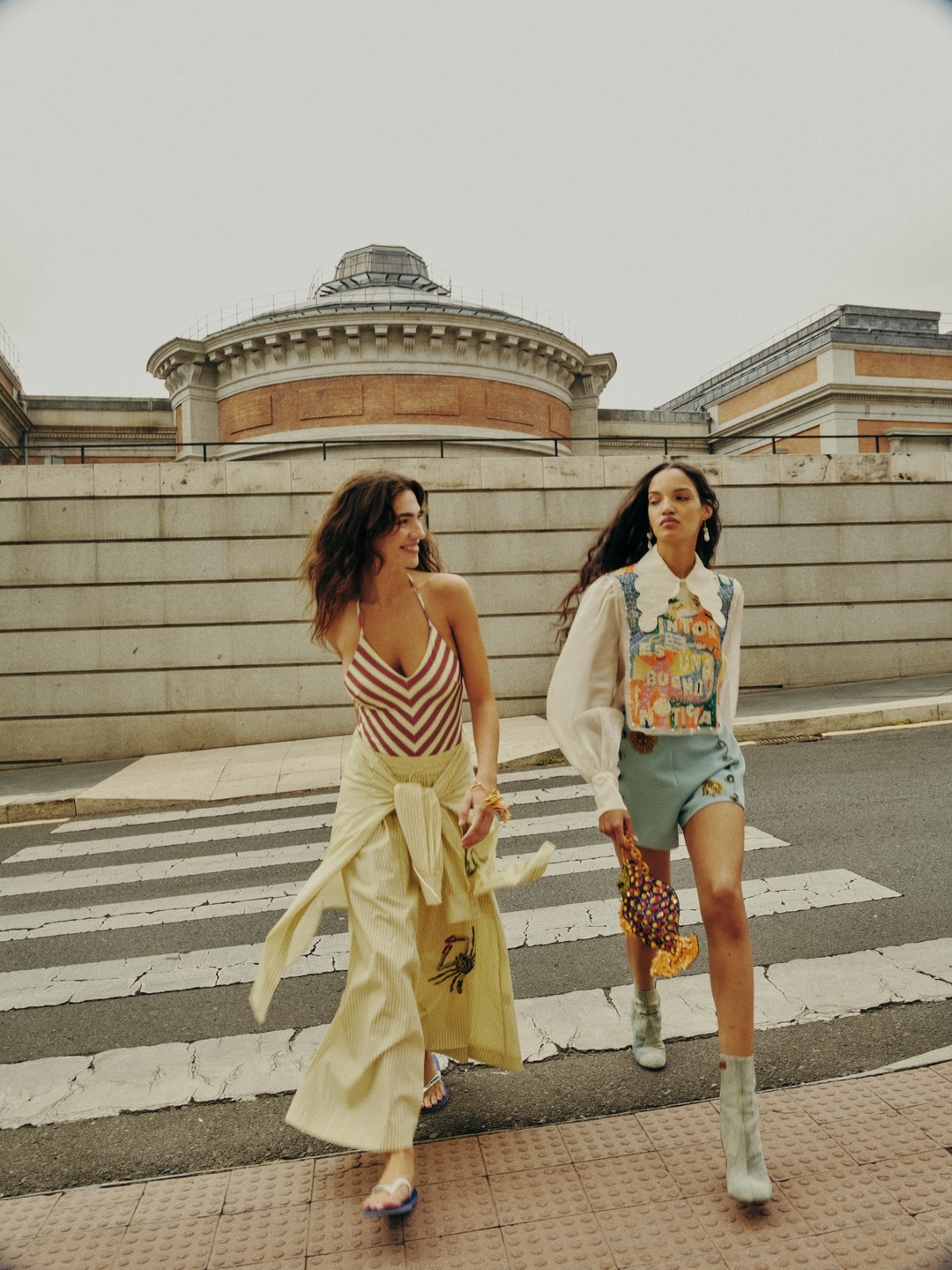 Two women in fashionable outfits walk across a crosswalk with an ornate stone building in the background, captured in an outdoor shot.