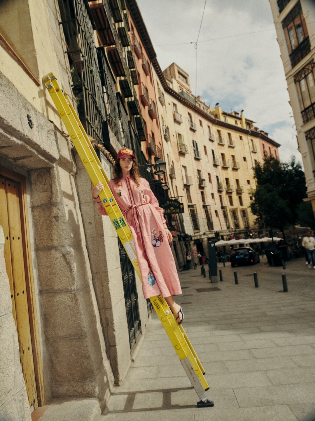 Woman in pink dress and matching baseball cap poses on yellow ladder against apartment building in city street in outdoor fashion shot.
