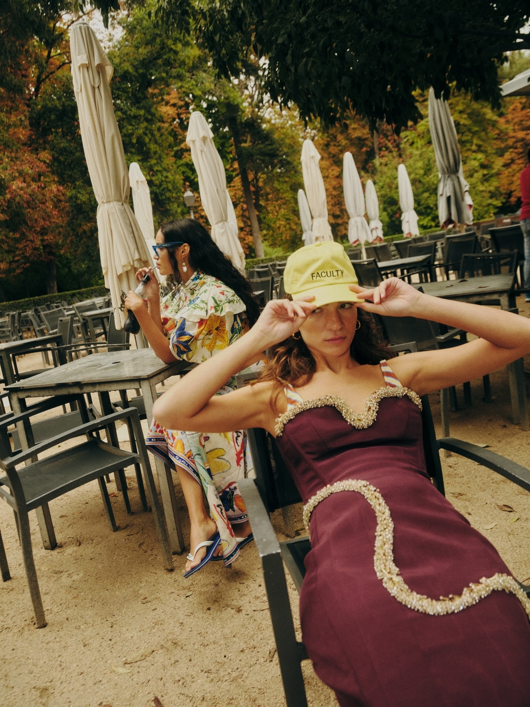 Two women in dresses sit in a sunny outdoor park setting at metal tables. One wears a yellow baseball cap and a maroon dress.