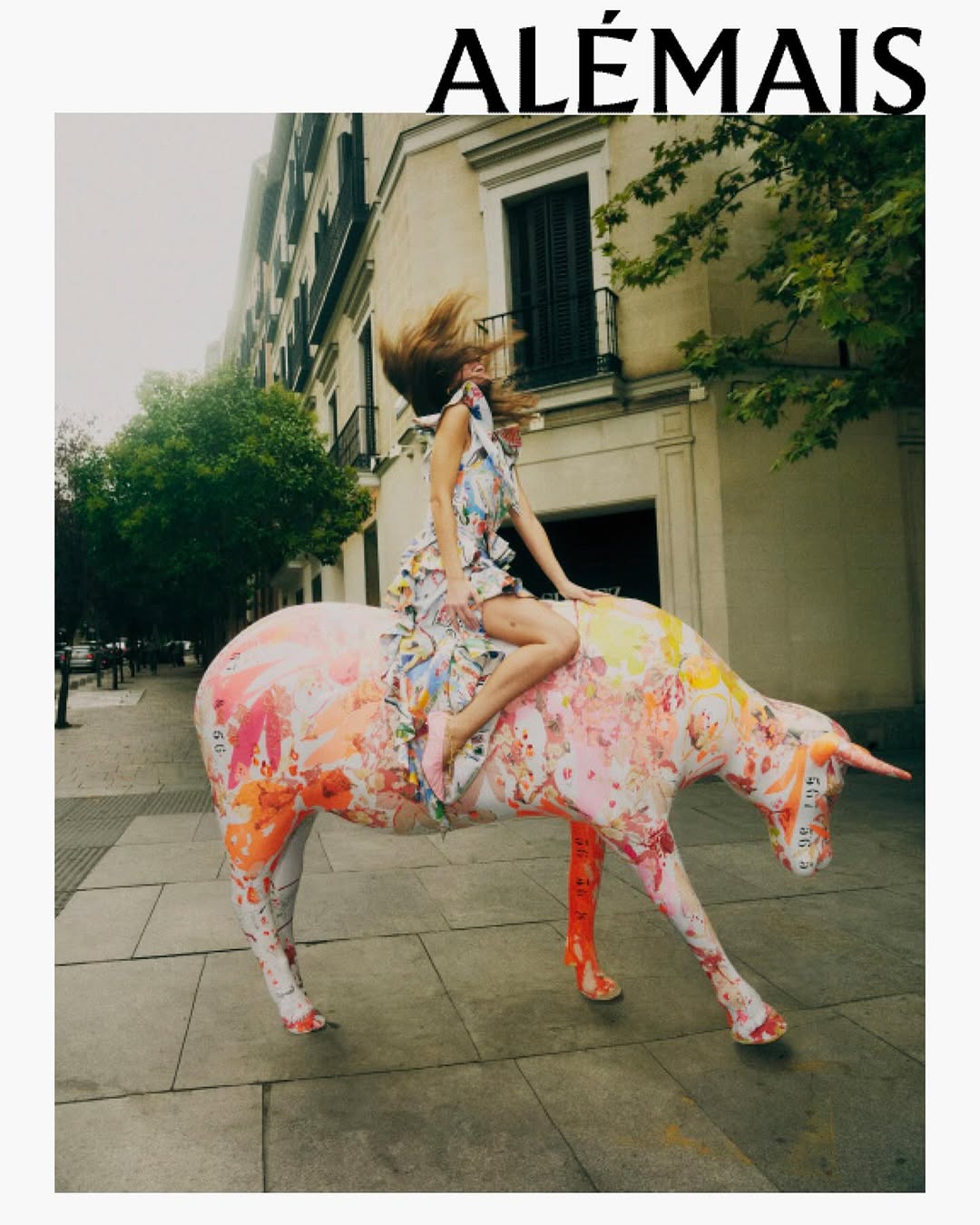An adult woman in a floral dress rides a colorful painted bull statue in an urban setting with buildings and trees in the background.