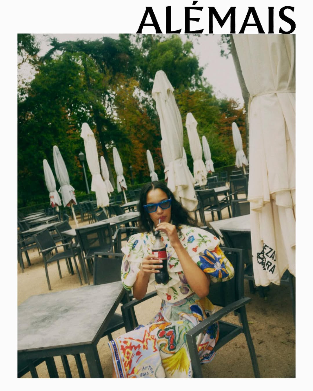 A woman in patterned clothing sitting at an outdoor cafe drinking from a Coca-Cola bottle with trees and umbrellas in the background.