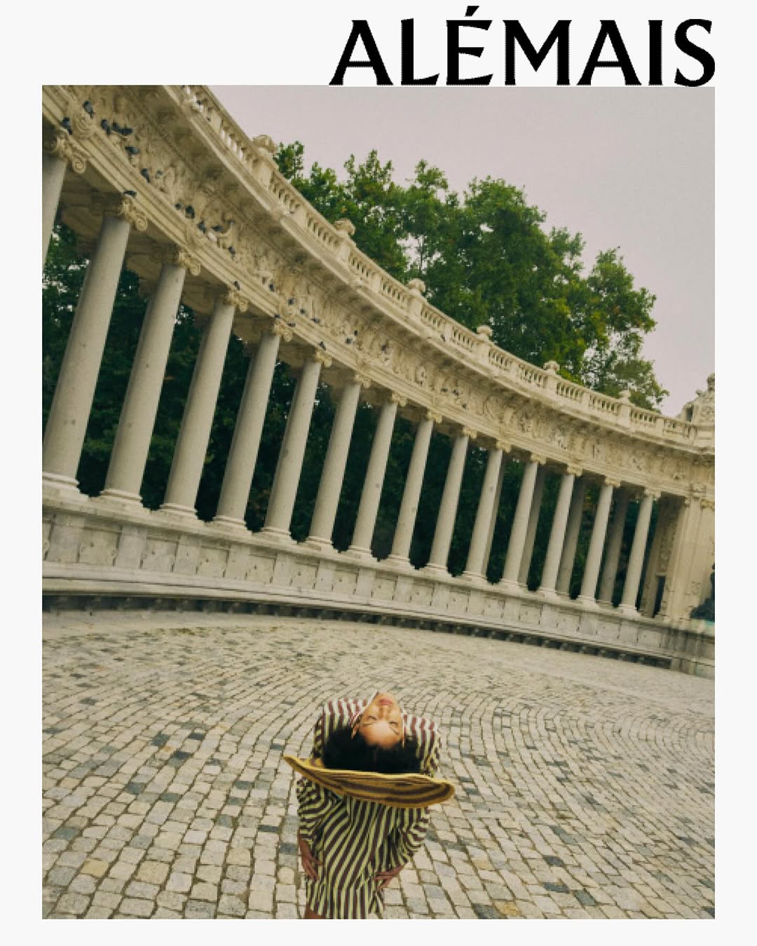 Woman in striped dress and hat standing on cobblestone, facing a marble colonnade with trees in the background, fashion outdoor shot.