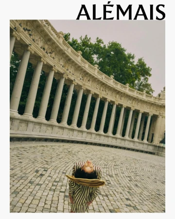 Woman in striped dress and hat standing on cobblestone, facing a marble colonnade with trees in the background, fashion outdoor shot.