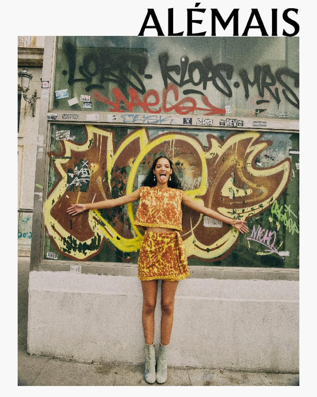 A young adult woman poses with her arms outstretched and tongue out while wearing a printed matching top and skirt against a graffiti wall.