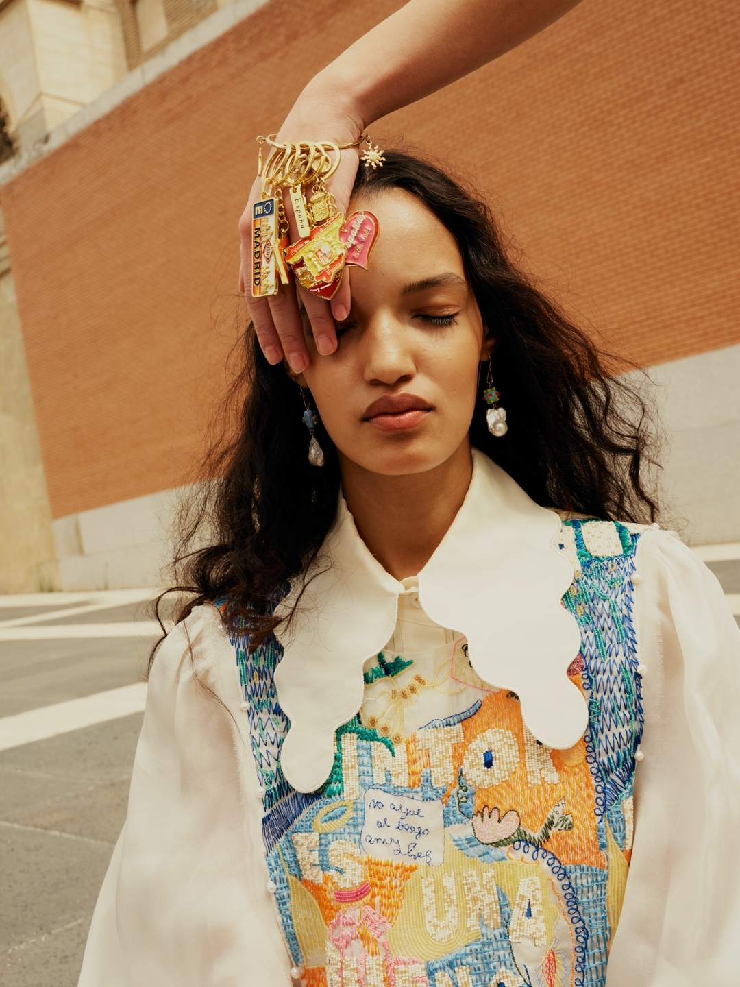 Woman in an outdoor setting with keychains on her hand, wearing earrings and a decorative top against a brick wall backdrop.