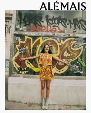 Model wearing a patterned yellow and brown two-piece outfit standing in front of a graffiti-covered storefront window.