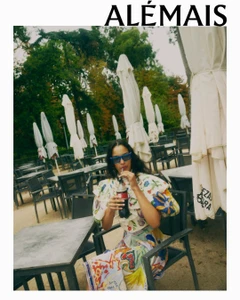 Woman in printed patterned blouse and sunglasses seated at an outdoor café table drinking from a bottle under white umbrellas
