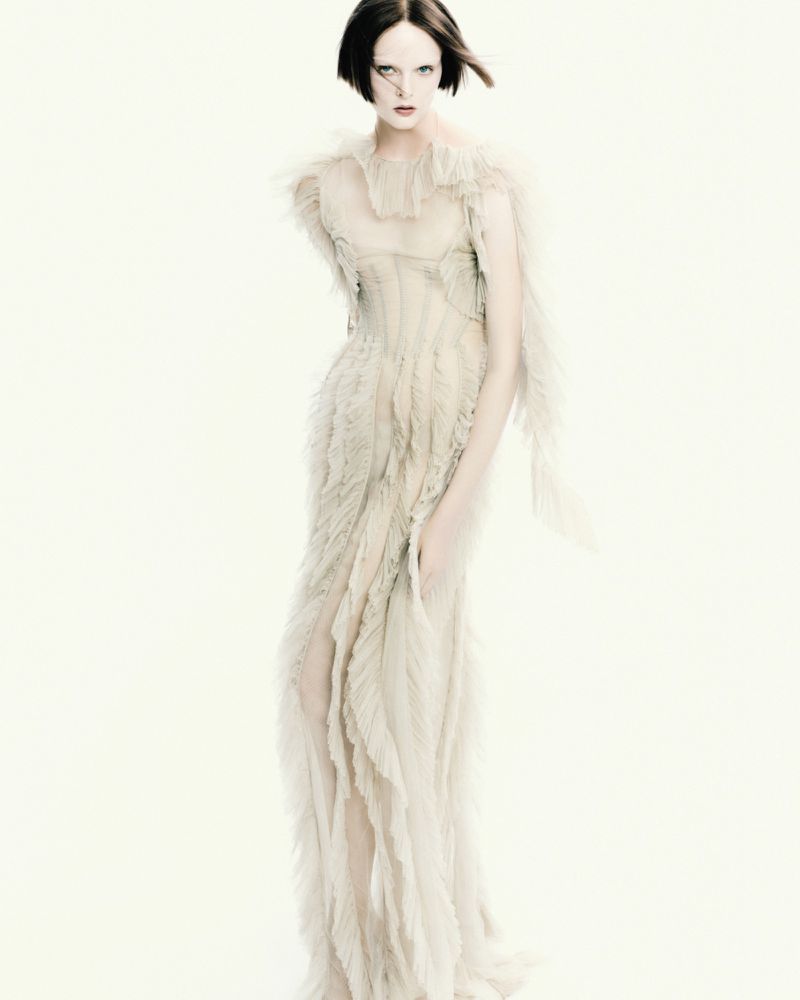 Fashion shot of a woman in a long beige dress posing against a bright white backdrop, featuring a muted color palette and artistic style.