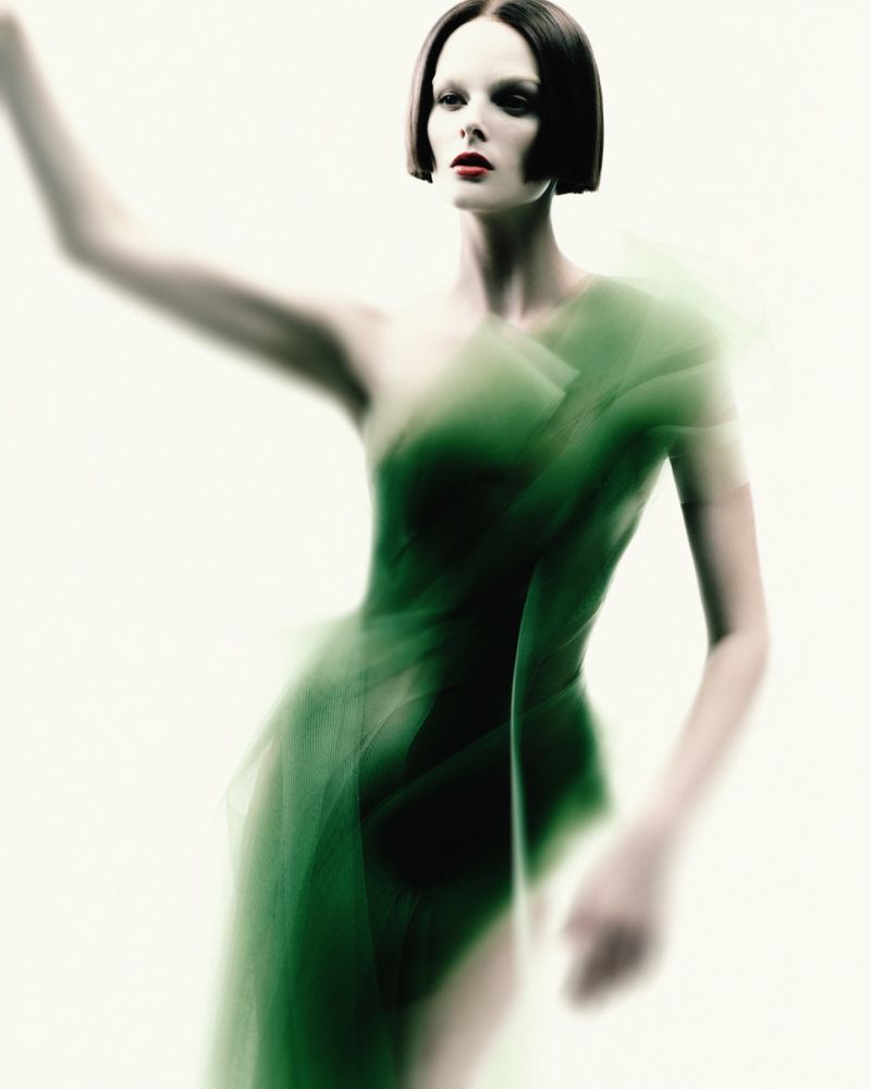 A woman in a green, translucent dress poses in a studio with an out-of-focus style, bright light and a sleek bob hairstyle.