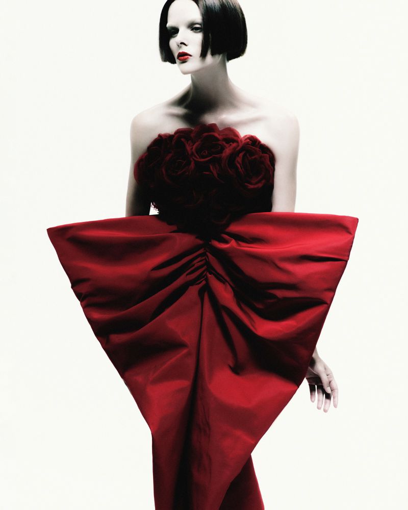 A fashion portrait of a woman with dark hair wearing a striking red dress with rose details and a large bow, set against a white background.