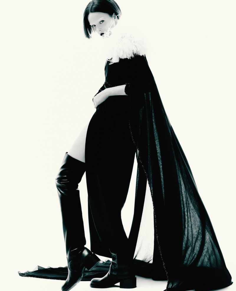 Fashionable woman poses in a black dress with a furry collar, black cape, and thigh-high leather boots against a bright white background.