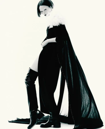 Fashionable woman poses in a black dress with a furry collar, black cape, and thigh-high leather boots against a bright white background.