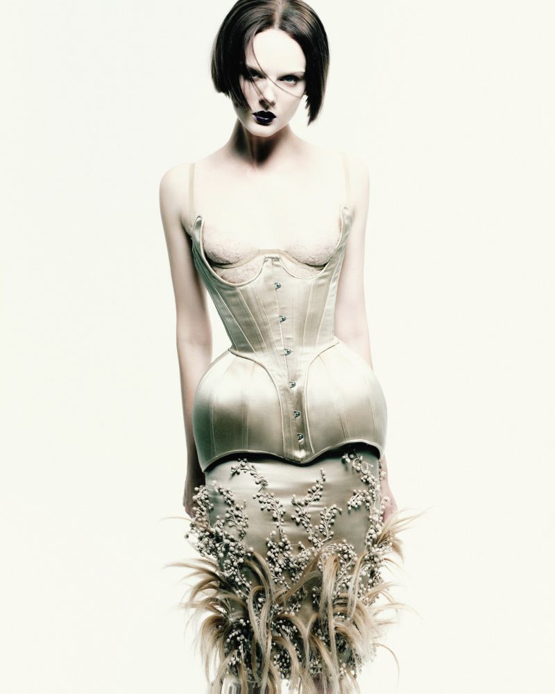 A striking fashion shot featuring a pale-skinned adult female model in a decorative corset and skirt against a bright white backdrop.