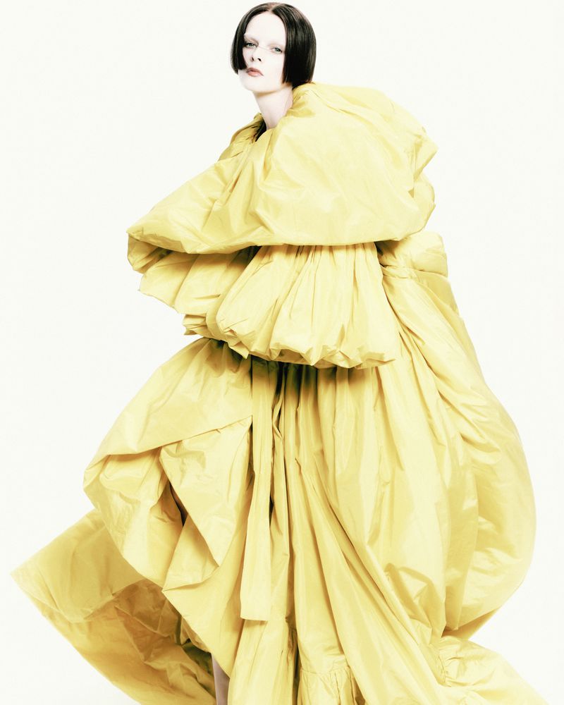 Fashionable person in an oversized, yellow gown with large ruffles, posing in a photo studio with a bright white background. High-fashion aesthetic.