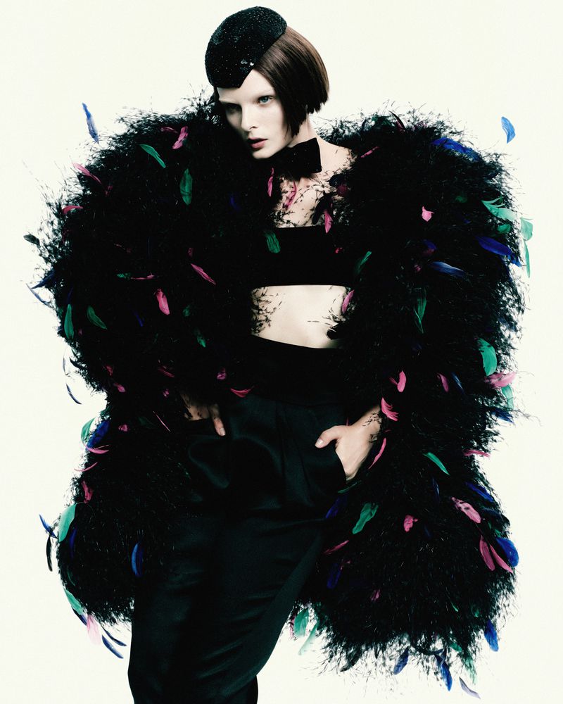 Fashion portrait of a model in a black feathered coat, strapless top, hat, and bow tie against a white background in a studio.