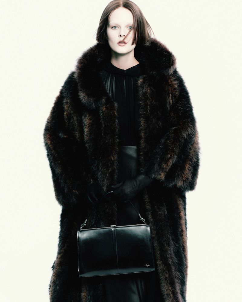 Model wearing a voluminous dark fur coat and black gloves holding a leather handbag against a plain white background