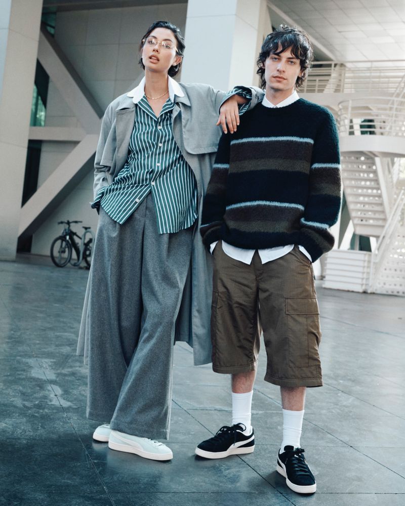 Two fashion models stand together outdoors against a modern building; the woman wears a striped shirt, and the man wears a striped sweater.