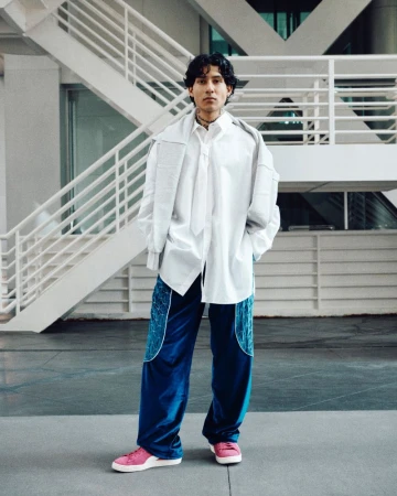 Man in an oversized white shirt and blue pants standing against a white and grey architectural background, wearing pink shoes.