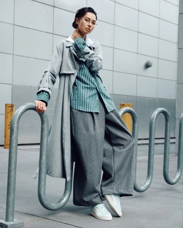 A stylish young woman in a grey trench coat and wide-leg trousers poses against an urban wall, leaning against a metal bike rack.