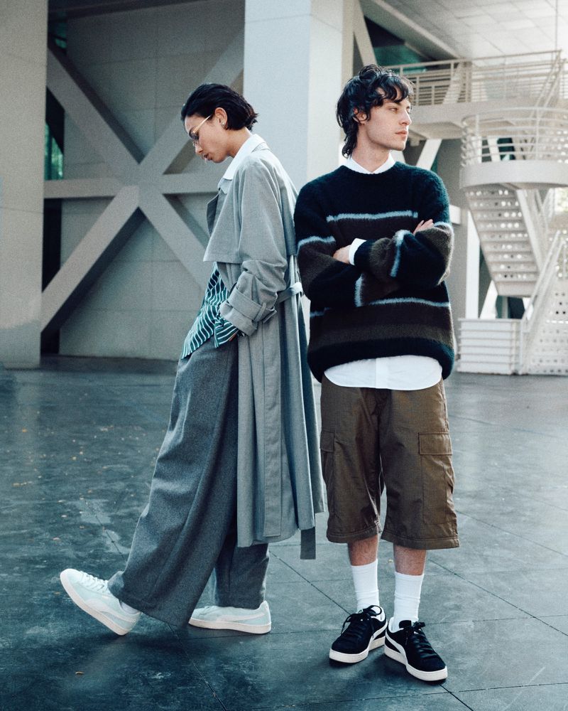 Two men in contemporary streetwear pose in a neutral indoor space with modern architectural details, showcasing current fashion trends.