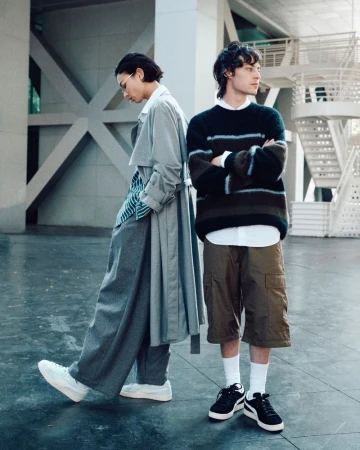 Two men in contemporary streetwear pose in a neutral indoor space with modern architectural details, showcasing current fashion trends.