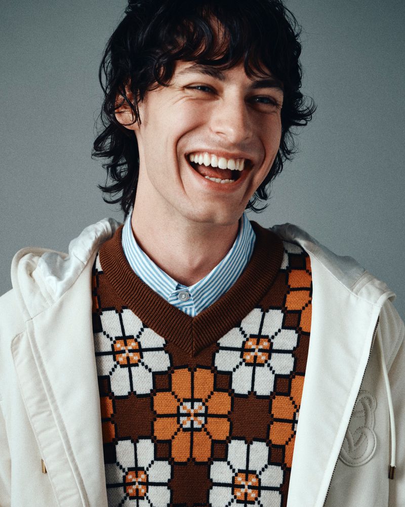 Smiling man with wavy dark hair wearing a patterned floral knit vest over a striped collared shirt and off-white hoodie.