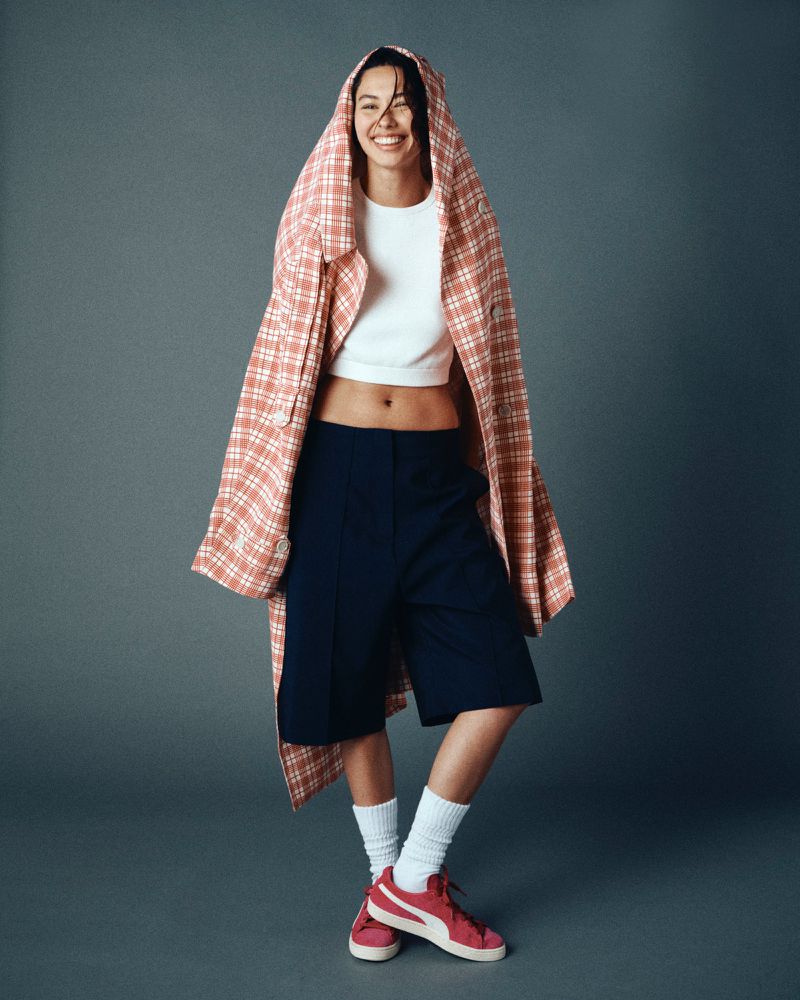 Smiling woman in white top, blue shorts, and pink sneakers stands against a grey backdrop while draped in a pink plaid jacket.