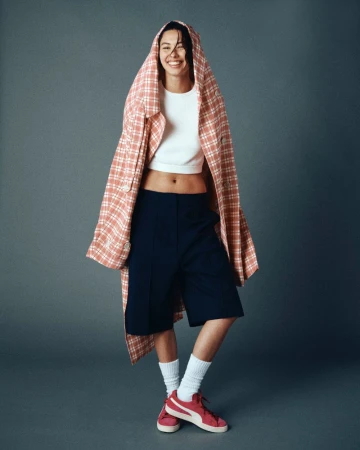 Smiling woman in white top, blue shorts, and pink sneakers stands against a grey backdrop while draped in a pink plaid jacket.