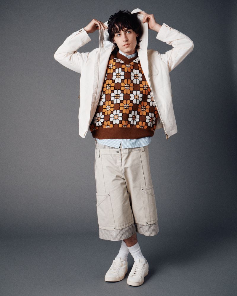 Male model poses in a hooded jacket, floral sweater, and khaki shorts against a gray backdrop, showcasing casual fashion.