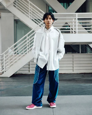 Man wearing a white shirt, blue pants, and pink sneakers posing in front of modern staircases for Office Magazine.