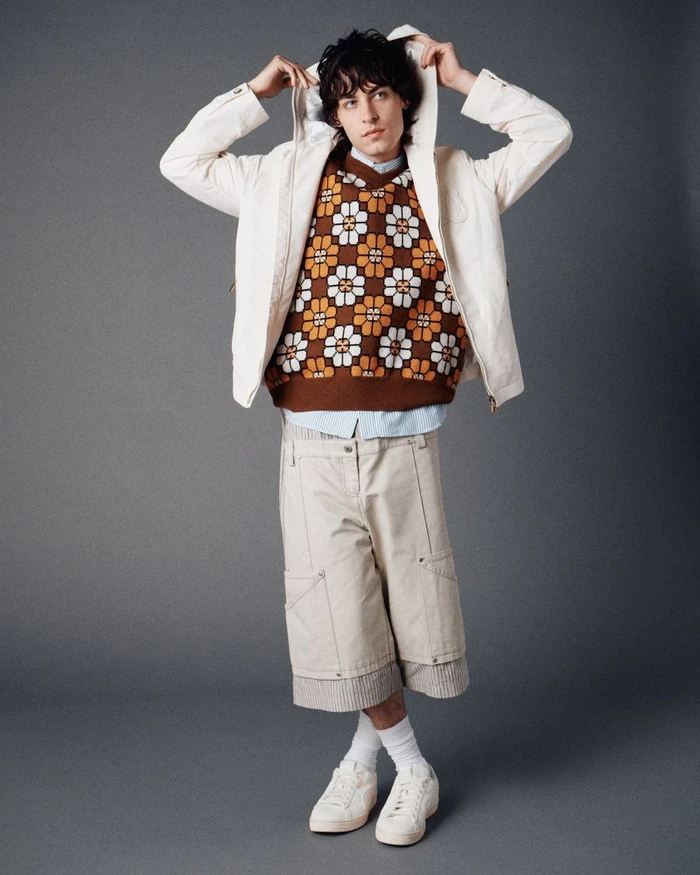 Model wearing a brown floral knit sweater vest, cream hooded jacket, beige wide shorts, and white sneakers against a grey backdrop.