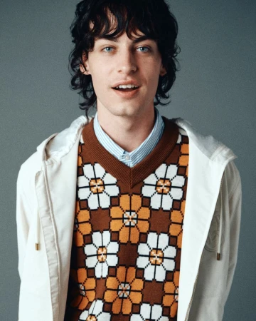 Portrait of a man with dark curly hair wearing a floral geometric sweater vest and white hoodie against a grey backdrop.