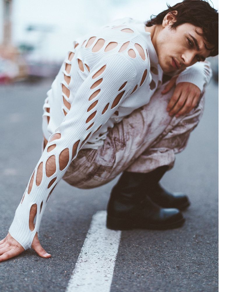 Man crouches on asphalt in a white cut-out sweater and patterned pants with black boots, creating a fashion-forward editorial image.