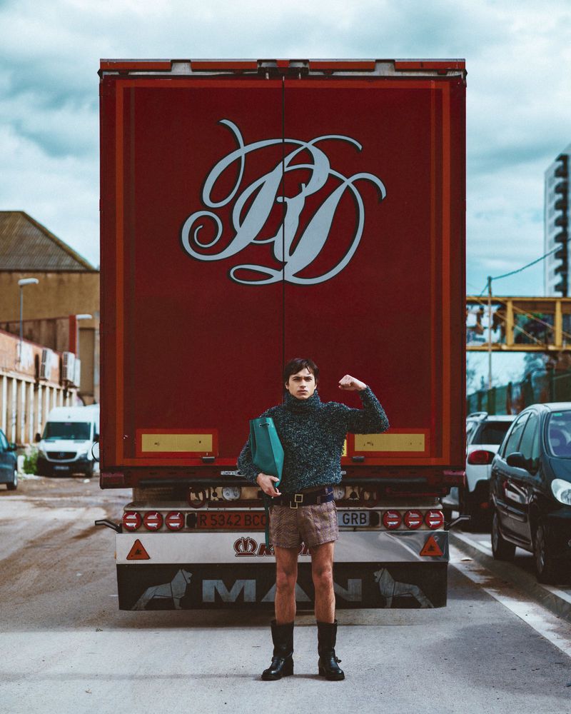 Young adult stands in front of a red semi-truck wearing a grey sweater, brown shorts and black boots in an urban setting.