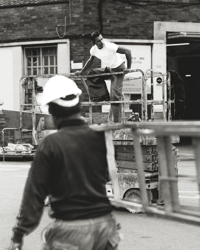 Two construction workers at an outdoor urban work site, one on a lift, one on the ground carrying a ladder in black and white.