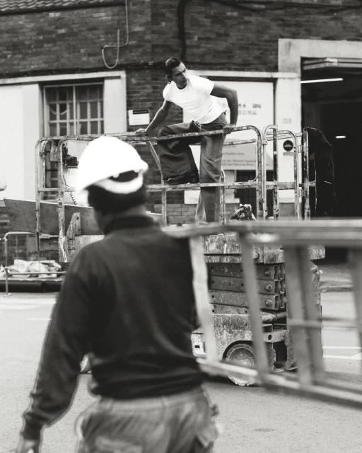 Two construction workers at an outdoor urban work site, one on a lift, one on the ground carrying a ladder in black and white.