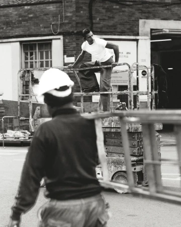 Two construction workers at an outdoor urban work site, one on a lift, one on the ground carrying a ladder in black and white.