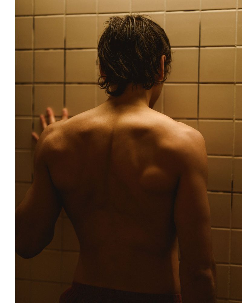 Back view of a light-skinned adult man leaning against a tiled wall indoors, lit in warm tones, showing his back and shoulders.